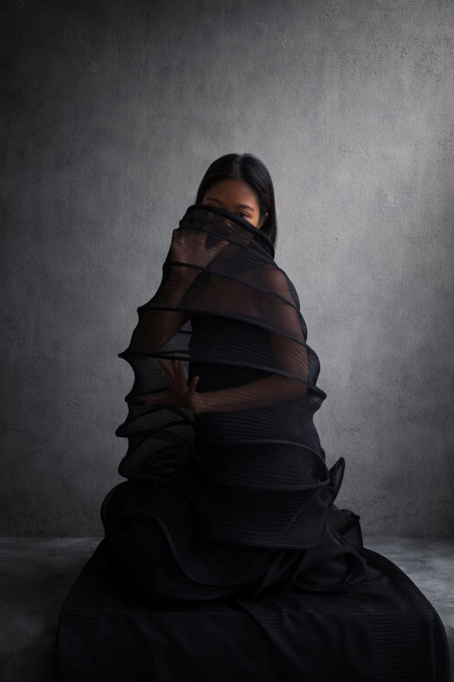 Figure wrapped in spiralling black pleated mesh against concrete